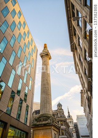 A stone column reaches towards the sky, surrounded by modern buildings. This is the Monument to the Great Fire of London, a historic site located in the heart of London. 137382808