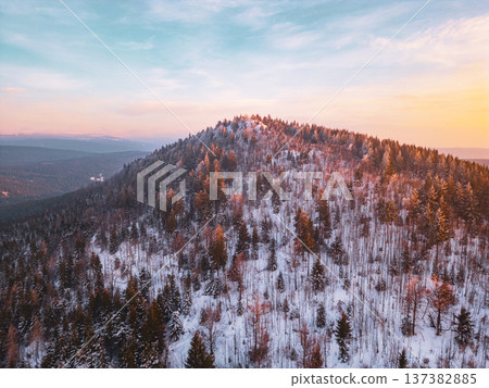 A drone captures Bukovec Mountain during sunset in the Jizera Mountains. The snow-covered terrain showcases trees on the slopes with fading light creating a colorful sky. 137382885