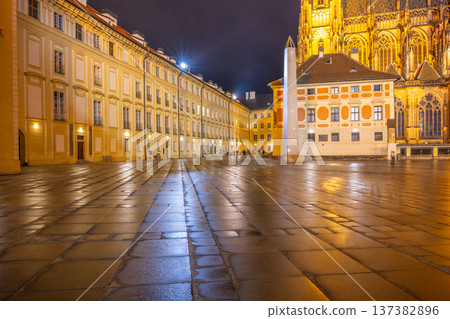 The Mrakotin Monolith stands tall at Prague Castle, surrounded by historical buildings, as warm light casts dramatic shadows on the cobblestone ground under the night sky. 137382896