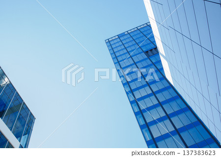 Modern skyscraper building exterior facade under clear blue sky low angle view. Minimalistic repetitive windows made of reflective glass. Financial district buildings, contemporary architecture. Modern skyscraper building exterior facade under clear blue sky low angle view. Minimalistic repetitive windows made of reflective glass. Financial district buildings, contemporary architecture. 137383623