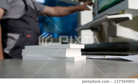 Close-up of white wooden boards on a workbench, with a factory worker operating machinery in the background. 137384903