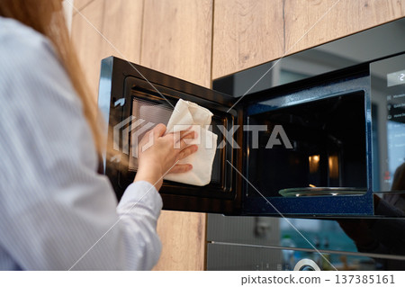 Woman wipes inner mesh surface of open built in microwave oven door with cloth in kitchen cabinet Woman wipes inner mesh surface of open built in microwave oven door with cloth in kitchen cabinet 137385161