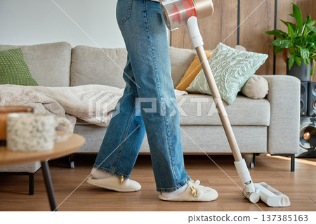 Woman in jeans using cordless vacuum cleaner on living room floor near sofa Woman in jeans using cordless vacuum cleaner on living room floor near sofa 137385163
