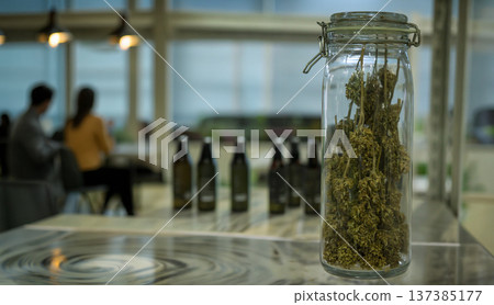 Dried cannabis buds in a clear glass jar on a counter at a modern dispensary or cafe, with people sitting in a blurred background. Perfect for lifestyle and retail themes Dried cannabis buds in a clear glass jar on a counter at a modern dispensary or cafe, with people sitting in a blurred background. Perfect for lifestyle and retail themes 137385177