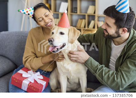 Happy couple celebrating dog birthday at home, party hats and gift on sofa 137385706