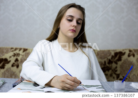 Focus on hand and blurred image of woman in creative mood while painting. Cozy home environment for relaxing while painting. Project concept for painting and creative leisure at home 137386107