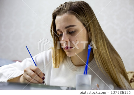 Young woman with long hair carefully paints with brush while sitting at table. Jar of water with brushes is visible next to her, soft background emphasizes true moment of painting at home Young woman with long hair carefully paints with brush while sitting at table. Jar of water with brushes is visible next to her, soft background emphasizes true moment of painting at home 137386135