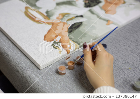 Close up of hand applying paint to brush in prepared jar with number written on it. Applying paint to corresponding area on canvas of paint by numbers painting Close up of hand applying paint to brush in prepared jar with number written on it. Applying paint to corresponding area on canvas of paint by numbers painting 137386147