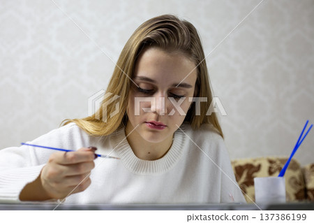 Young woman with long blonde hair focuses on painting with small brush on canvas while seated at table, surrounded by art supplies and decorative loft background in bright indoor setting Young woman with long blonde hair focuses on painting with small brush on canvas while seated at table, surrounded by art supplies and decorative loft background in bright indoor setting 137386199