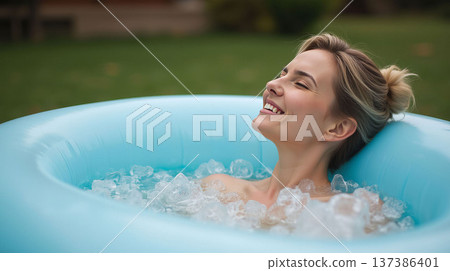 A woman relaxing in an inflatable tub filled with ice cubes, smiling with joy, outdoors in a lush green garden. Wellness and relaxation. Inflatable Ice Bath Concept For Home 137386401