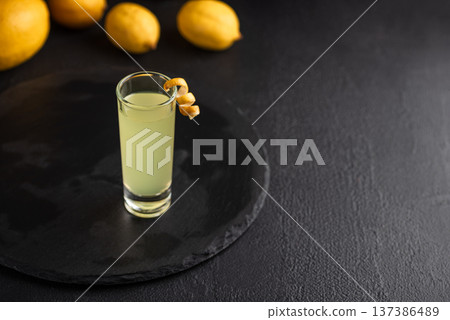 Tall shot glass of limoncello garnished with a curled lemon peel, surrounded by fresh lemons on dark textured background 137386489