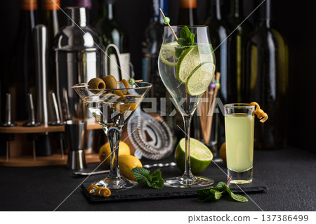 Bar composition featuring a variety of cocktails: martini with olives, citrus mint highball, and limoncello shot against wine bottles, shaker, strainer and fresh ingredients 137386499