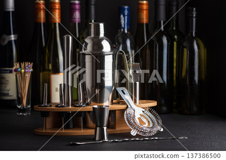 Professional cocktail bar composition featuring a stainless steel shaker, jigger, strainer, muddler, zester, and pour spouts neatly arranged on a wooden stand against wine bottles in background 137386500