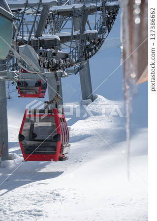 Red cable car gondola moving along ski lift line with snow covered mountain slope and icicles in winter resort landscape. Winter tourism marketing, ski resort promotion, mountain transport Red cable car gondola moving along ski lift line with snow covered mountain slope and icicles in winter resort landscape. Winter tourism marketing, ski resort promotion, mountain transport 137386662