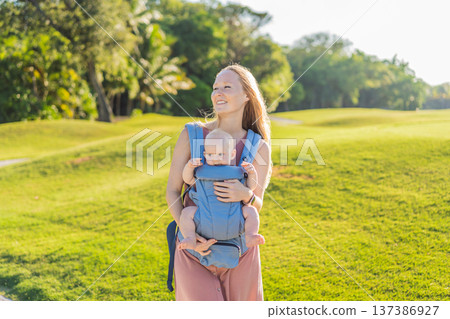 Mother using ergonomic baby carrier during a walk with her infant outdoors. Comfortable babywearing, active motherhood lifestyle, bonding, modern parenting, hands free care and everyday family routine Mother using ergonomic baby carrier during a walk with her infant outdoors. Comfortable babywearing, active motherhood lifestyle, bonding, modern parenting, hands free care and everyday family routine 137386927