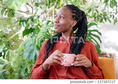 Dreaming african american woman holding cup resting on armchair surround houseplants looking aside. Dreaming african american woman holding cup resting on armchair surround houseplants looking aside. 137387217