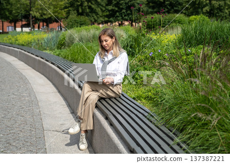 Positive businesswoman working on remote in park, sitting on bench talking by video call on laptop. Positive businesswoman working on remote in park, sitting on bench talking by video call on laptop. 137387221