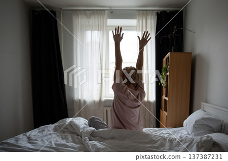 Relaxed middle aged woman stretching arms and body sitting in bed after waking up in morning. Relaxed middle aged woman stretching arms and body sitting in bed after waking up in morning. 137387231