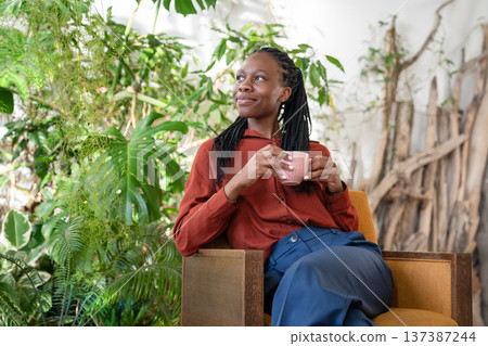 Dreaming african american woman holding cup resting on armchair surround houseplants looking aside. 137387244