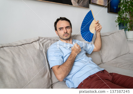 Sad man suffering from heat in apartment, fanning himself with paper fan, no air conditioner at home 137387245