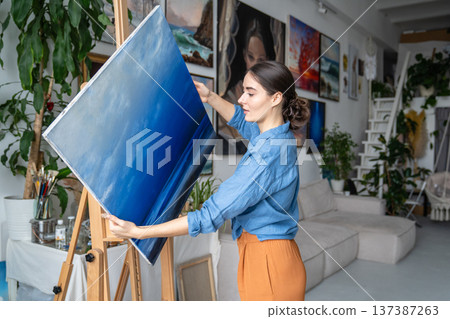 Professional female artist painter carefully taking off canvas from easel. holding finished picture. Professional female artist painter carefully taking off canvas from easel. holding finished picture. 137387263