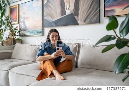 Curious woman artist sitting on couch watching media online on smartphone at cozy home gallery. Curious woman artist sitting on couch watching media online on smartphone at cozy home gallery. 137387271