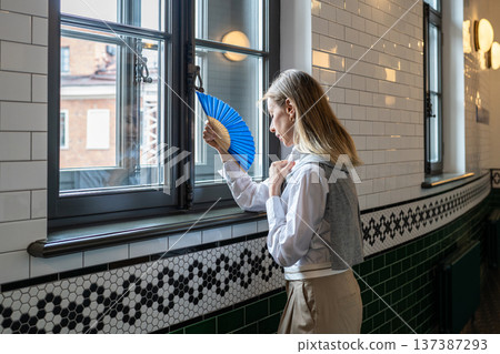 Woman feeling suffering from heatstroke and dizziness indoors, waving paper fan in hand to cool down Woman feeling suffering from heatstroke and dizziness indoors, waving paper fan in hand to cool down 137387293