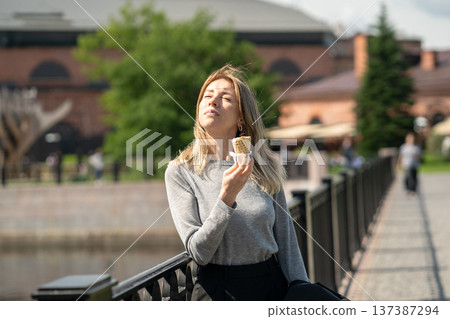 Carefree business woman enjoys sunny weather and ice cream, stands on bridge squinting at bright sun 137387294