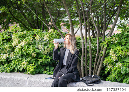 Relaxed businesswoman rests after day in office in park enjoys weather, drinking water from bottle. 137387304
