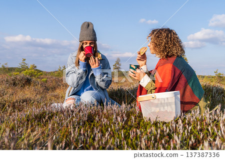 Couple of relaxing sisters holds picnic sitting on meadow grass on sunny autumn day Couple of relaxing sisters holds picnic sitting on meadow grass on sunny autumn day 137387336