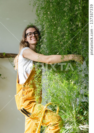 Joyful young woman gardener standing on stepladder, embracing lush asparagus fern houseplant. 137387339