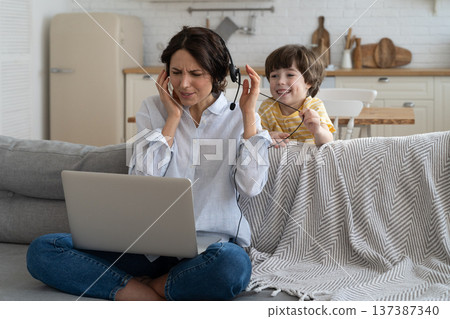 Exhausted mother sitting on couch at home, working on laptop, child distracted and asking attention Exhausted mother sitting on couch at home, working on laptop, child distracted and asking attention 137387340