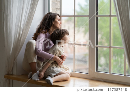 Mom and curious kid look outside sit on window sill. Mother or babysitter exploring world from home 137387348