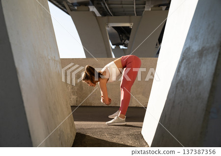 Young female jogger prepare for running workout, stretch legs, bend forward outdoors under bridge 137387356