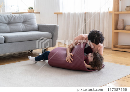 Mother wrapped smiling kid in yoga mat playing with son after sport exercise at home during lockdown Mother wrapped smiling kid in yoga mat playing with son after sport exercise at home during lockdown 137387366