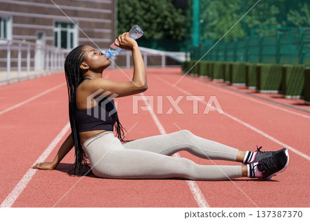 African American woman sitting in stadium drinking a lot of water from bottle after intense workout. African American woman sitting in stadium drinking a lot of water from bottle after intense workout. 137387370