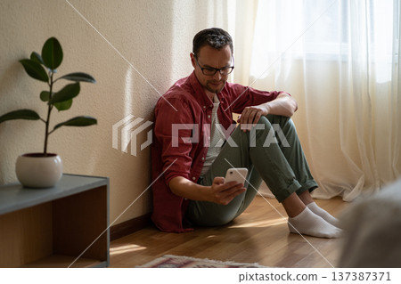 Upset young man holding smartphone waiting for ex-girlfriend call while sitting on floor at home Upset young man holding smartphone waiting for ex-girlfriend call while sitting on floor at home 137387371
