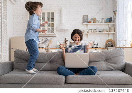Tired mother sit on couch, work on laptop at home, hyperactive little kid jumping drawing attention. Tired mother sit on couch, work on laptop at home, hyperactive little kid jumping drawing attention. 137387448