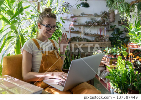 Woman gardener using laptop smiling talking in video chat say hello her friend sitting in greenhouse 137387449