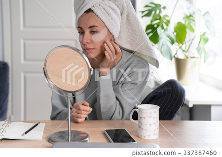 Woman with towel on head apply hydrogel under-eye recovery patches at home, looking in mirror. Woman with towel on head apply hydrogel under-eye recovery patches at home, looking in mirror. 137387460