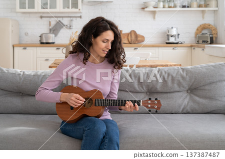 Woman recording content to vlog, playing on ukulele guitar looking at webcam during lockdown at home 137387487