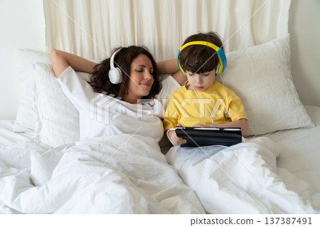 Family morning in bedroom: mom in headphones look at little kid son play games on tablet computer 137387491