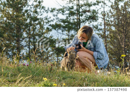 Woman photographer plays with lonely cat walking on forest meadow against blurry green trees 137387498