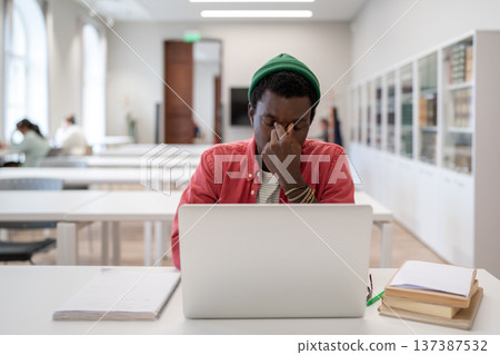 Tired black student guy massaging nose sitting with closed eyes in front of laptop in library 137387532