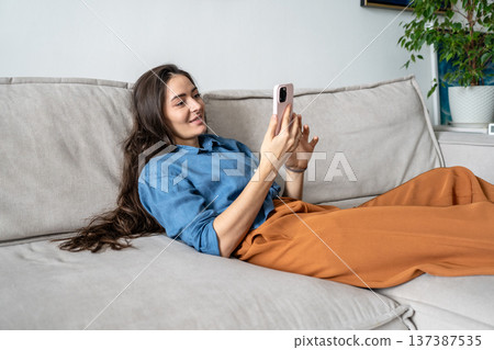 Smiling positive woman artist lying on couch scrolling social media online on smartphone at home. 137387535
