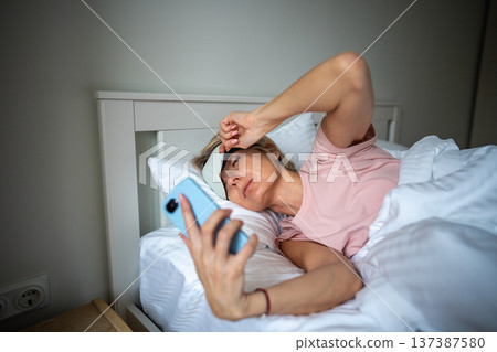 Sleepy woman peeking out from eye mask and checking time on smartphone, lazy early morning in bed. Sleepy woman peeking out from eye mask and checking time on smartphone, lazy early morning in bed. 137387580