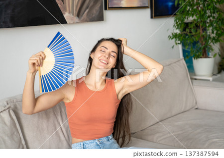 Woman at home coping with overheating during summer season, sitting on sofa and waving hand fan. 137387594
