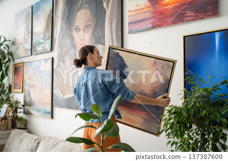Focused skillful woman artist hanging painting on wall filled with exhibition of own masterpieces. Focused skillful woman artist hanging painting on wall filled with exhibition of own masterpieces. 137387600