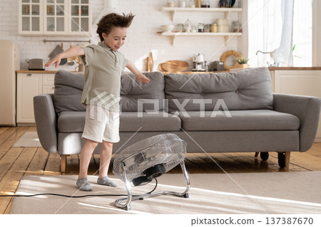 Little boy enjoy air flow from fan in living room. Cute small kid play with ventilator blowing wind Little boy enjoy air flow from fan in living room. Cute small kid play with ventilator blowing wind 137387670