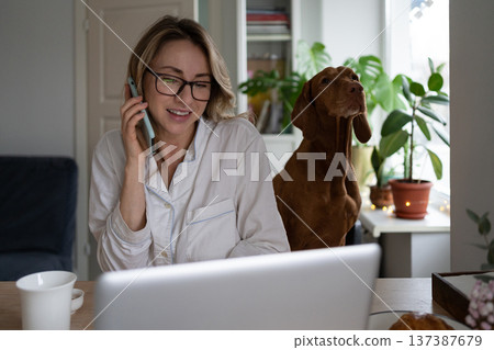 Woman in pajamas sitting with dog in living room, taking on mobile phone, remote working at laptop Woman in pajamas sitting with dog in living room, taking on mobile phone, remote working at laptop 137387679
